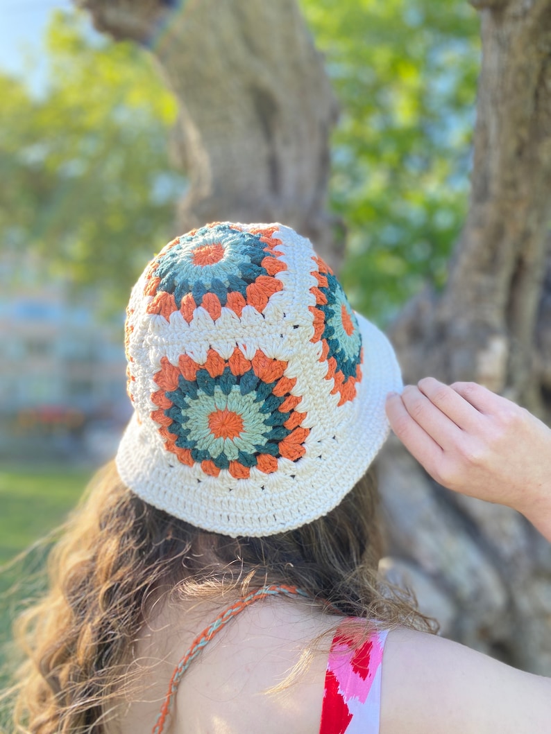 Puede incluir: Un sombrero de cubo de crochet blanco con un patr&oacute;n geom&eacute;trico colorido en tonos naranja, verde y blanco.