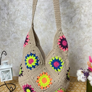 Op de afbeelding: Een beige gehaakte tote bag met een kleurrijk bloemenpatroon. De tas heeft een lange band en is gemaakt van een zacht, getextureerd materiaal.