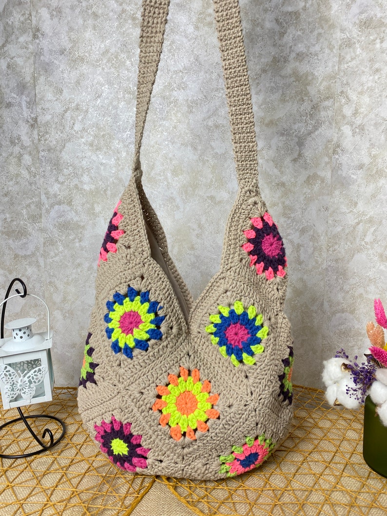 Op de afbeelding: Een beige gehaakte tote bag met een kleurrijk bloemenpatroon. De tas heeft een lange band en is gemaakt van een zacht, getextureerd materiaal.
