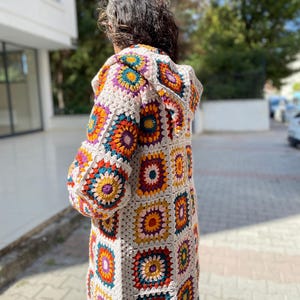 Puede incluir: C&aacute;rdigan largo con capucha de crochet, con un patr&oacute;n de cuadrados de la abuela. El c&aacute;rdigan presenta un color base neutro con cuadrados coloridos en tonos naranja, amarillo, morado, turquesa y blanco. Las mangas son largas y el c&aacute;rdigan llega a la mitad del muslo.