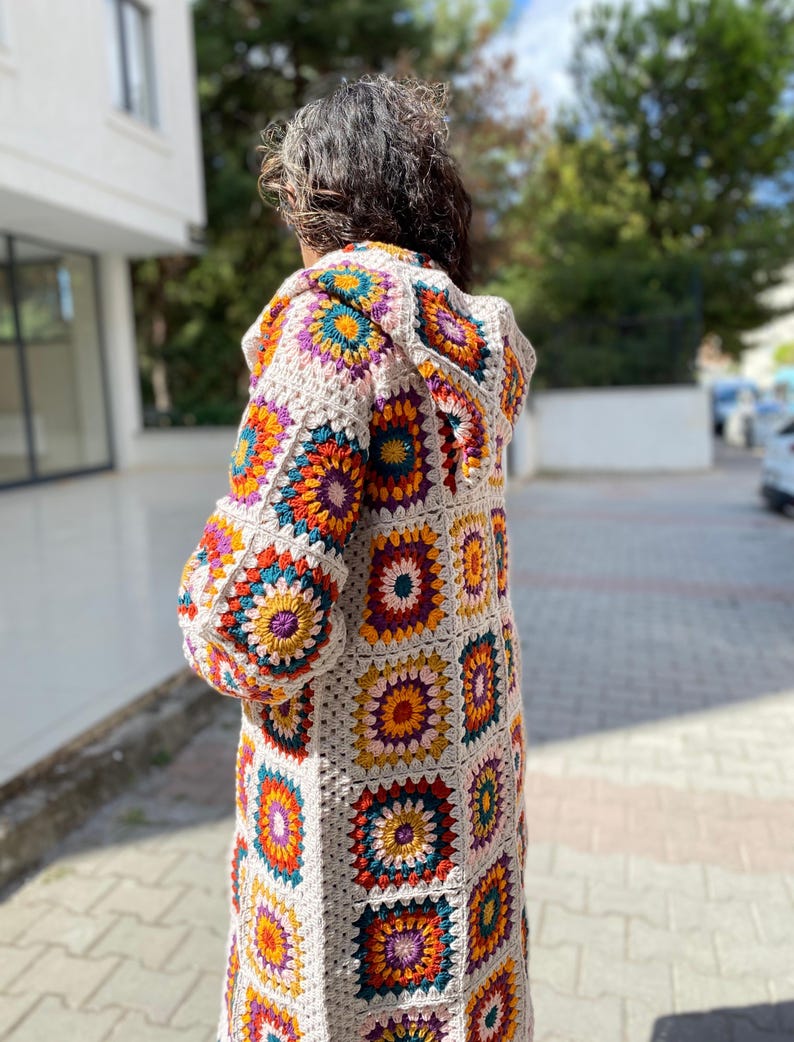 Puede incluir: C&aacute;rdigan largo con capucha de crochet, con un patr&oacute;n de cuadrados de la abuela. El c&aacute;rdigan presenta un color base neutro con cuadrados coloridos en tonos naranja, amarillo, morado, turquesa y blanco. Las mangas son largas y el c&aacute;rdigan llega a la mitad del muslo.