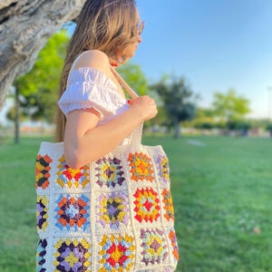 May include: A white crocheted tote bag with colorful granny squares. The bag has a long strap and is made with a variety of colors, including red, orange, yellow, green, blue, purple, and brown.
