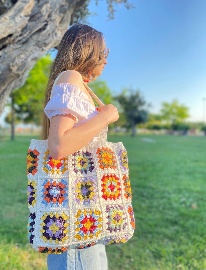 May include: A white crocheted tote bag with colorful granny squares. The bag has a long strap and is made with a variety of colors, including red, orange, yellow, green, blue, purple, and brown.