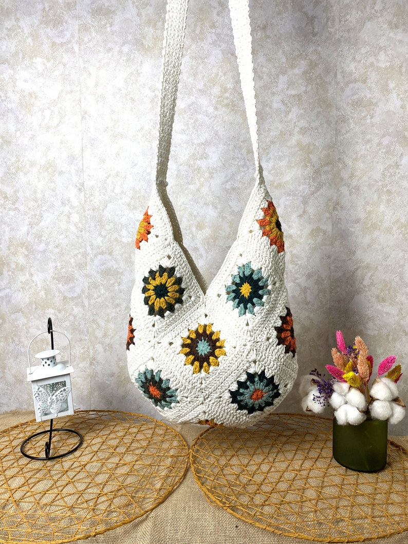 Op de afbeelding: Witte gehaakte tote bag met een geometrisch patroon van kleurrijke bloemen. De tas heeft een lange band en is gemaakt van een zacht, lichtgewicht materiaal.