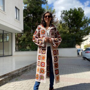 Puede incluir: Un abrigo largo con capucha de ganchillo con un dise&ntilde;o de patchwork de cuadrados crema, naranja, amarillo y marr&oacute;n. El abrigo se lleva sobre una camiseta blanca y unos vaqueros azules. La longitud del abrigo llega hasta los tobillos.