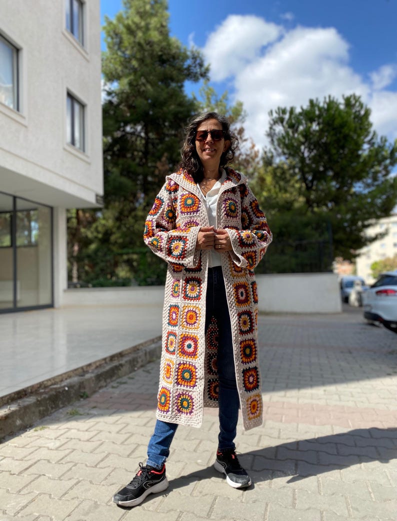 Puede incluir: Un abrigo largo con capucha de ganchillo con un dise&ntilde;o de patchwork de cuadrados crema, naranja, amarillo y marr&oacute;n. El abrigo se lleva sobre una camiseta blanca y unos vaqueros azules. La longitud del abrigo llega hasta los tobillos.
