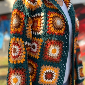 May include: A long, green crochet cardigan with a hood. The cardigan is made of granny squares with a multi-colored pattern of orange, brown, white, and yellow.