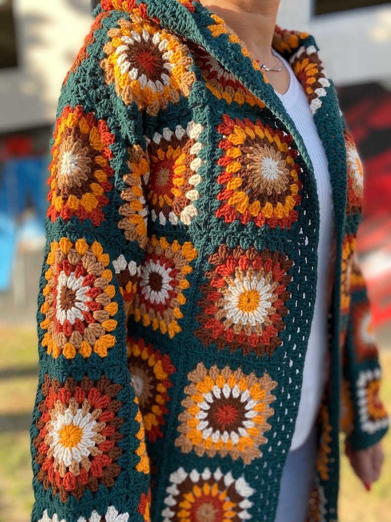 May include: A long, green crochet cardigan with a hood. The cardigan is made of granny squares with a multi-colored pattern of orange, brown, white, and yellow.