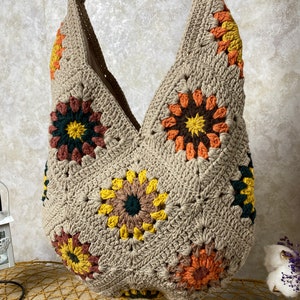 Op de afbeelding: Een beige gehaakte tote bag met een V-vormige bovenkant en kleurrijke bloemenontwerpen. De tas is gemaakt van dik garen en heeft een ontspannen, bohemian stijl.