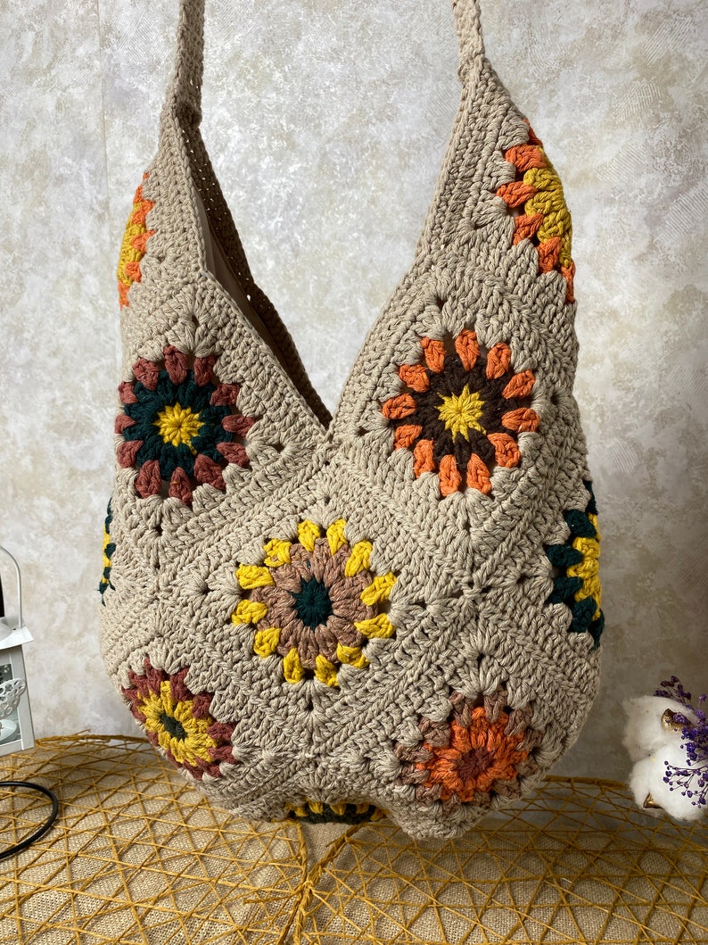 Op de afbeelding: Een beige gehaakte tote bag met een V-vormige bovenkant en kleurrijke bloemenontwerpen. De tas is gemaakt van dik garen en heeft een ontspannen, bohemian stijl.