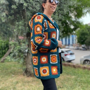 K&ouml;nnte beinhalten: Eine tealblaue geh&auml;kelte Strickjacke mit Kapuze, die ein farbenfrohes geometrisches Muster aus orangefarbenen, braunen, wei&szlig;en und cremefarbenen Quadraten aufweist.