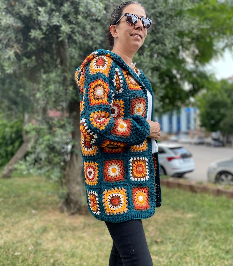 K&ouml;nnte beinhalten: Eine tealblaue geh&auml;kelte Strickjacke mit Kapuze, die ein farbenfrohes geometrisches Muster aus orangefarbenen, braunen, wei&szlig;en und cremefarbenen Quadraten aufweist.