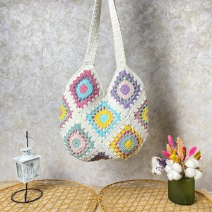 May include: A white crocheted tote bag with colorful floral designs. The bag has a long strap and is made of a soft, textured material.