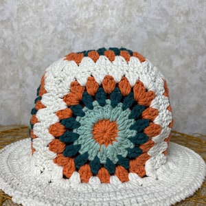 Puede incluir: Un sombrero de sol blanco de crochet con un patr&oacute;n geom&eacute;trico colorido en tonos naranja, verde y azul. El sombrero tiene un ala ancha y una corona redonda.