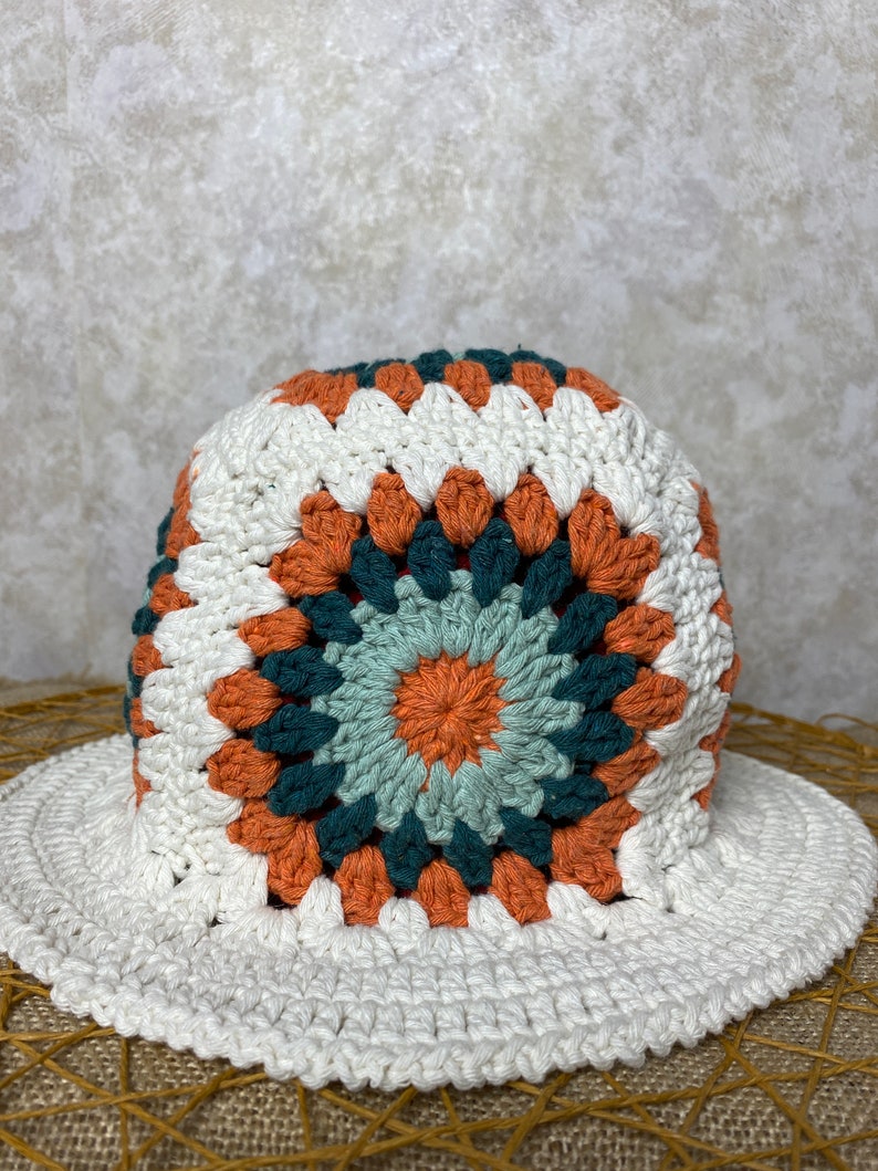 Puede incluir: Un sombrero de sol blanco de crochet con un patr&oacute;n geom&eacute;trico colorido en tonos naranja, verde y azul. El sombrero tiene un ala ancha y una corona redonda.