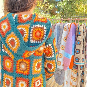 May include: A teal blue crocheted cardigan with a hood, featuring a colorful geometric pattern of orange, brown, and white. The cardigan is hanging on a rack with other crocheted garments.