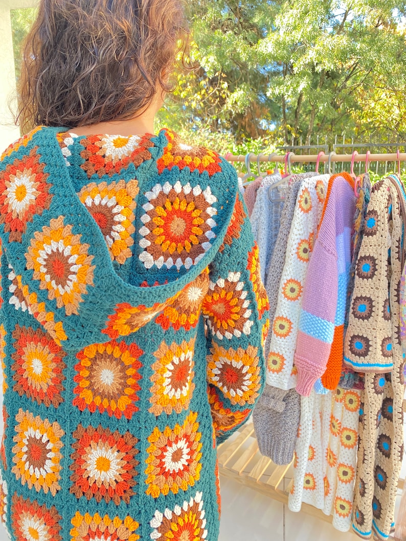 May include: A teal blue crocheted cardigan with a hood, featuring a colorful geometric pattern of orange, brown, and white. The cardigan is hanging on a rack with other crocheted garments.