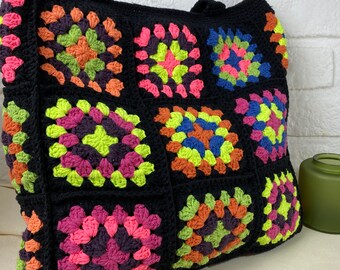 Peut inclure: Un sac cabas en crochet noir avec des carrés de grand-mère colorés. Les carrés présentent une variété de couleurs, notamment le rose, le vert, l'orange et le bleu.