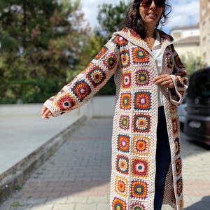 Puede incluir: Un abrigo largo de ganchillo color crema con capucha. El abrigo presenta patrones cuadrados coloridos en tonos naranja, amarillo, morado y verde. El abrigo est&aacute; abierto por delante y tiene mangas largas. El abrigo est&aacute; hecho a mano y parece ser de un estilo bohemio &uacute;nico.
