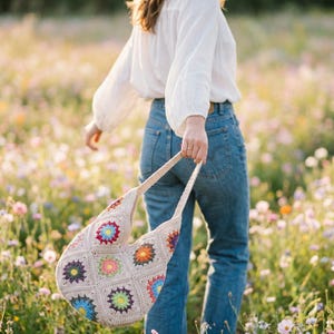 May include: A cream-coloured crochet shoulder bag with colourful floral squares. The bag is held by a person wearing blue jeans and a white blouse, walking through a field of wildflowers. The bag's design features red, blue, yellow, and green accents.
