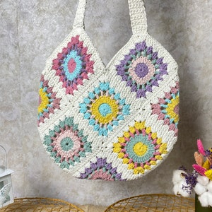 May include: A white crocheted tote bag with colorful granny square designs. The bag has a wide, rounded bottom and two long straps.