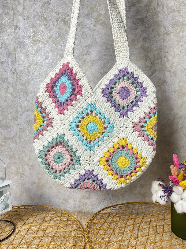 May include: A white crocheted tote bag with colorful granny square designs. The bag has a wide, rounded bottom and two long straps.