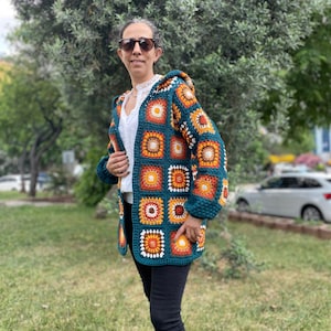 K&ouml;nnte beinhalten: Eine tealblaue geh&auml;kelte Strickjacke mit Kapuze. Die Strickjacke besteht aus Granny Squares mit orangefarbenen, gelben, wei&szlig;en und braunen Akzenten.