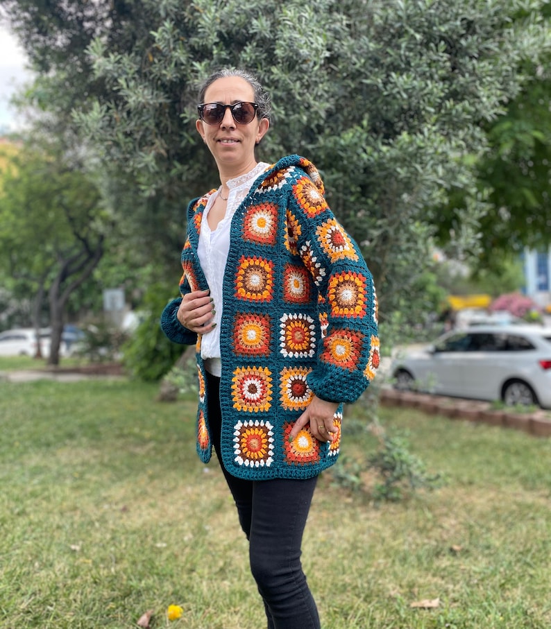 K&ouml;nnte beinhalten: Eine tealblaue geh&auml;kelte Strickjacke mit Kapuze. Die Strickjacke besteht aus Granny Squares mit orangefarbenen, gelben, wei&szlig;en und braunen Akzenten.