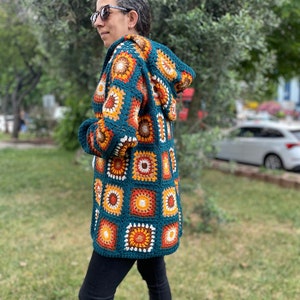 K&ouml;nnte beinhalten: Eine tealblaue geh&auml;kelte Strickjacke mit Kapuze, die ein farbenfrohes geometrisches Muster aus orangefarbenen, gelben und wei&szlig;en Quadraten aufweist.