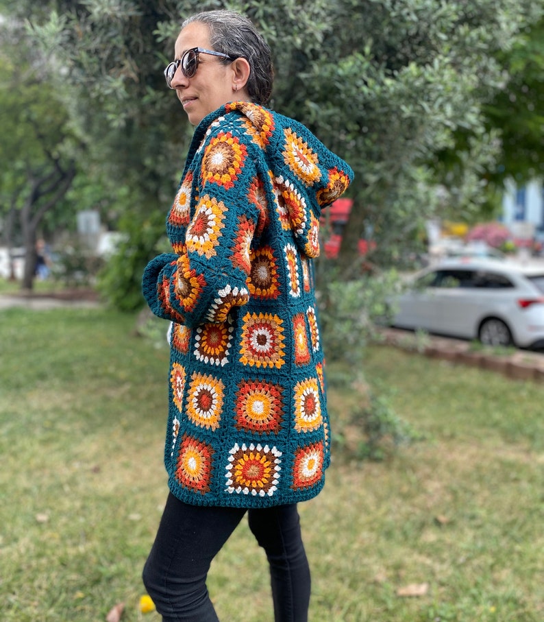 K&ouml;nnte beinhalten: Eine tealblaue geh&auml;kelte Strickjacke mit Kapuze, die ein farbenfrohes geometrisches Muster aus orangefarbenen, gelben und wei&szlig;en Quadraten aufweist.