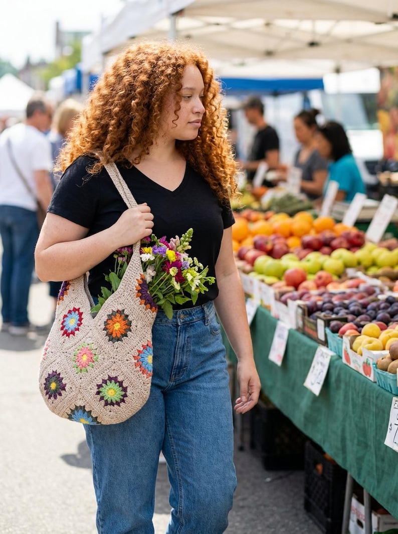 May include: A beige crochet tote bag with colourful floral designs, filled with a bouquet of fresh flowers. The bag is carried by a person wearing a black V-neck shirt and blue jeans, at an outdoor market.
