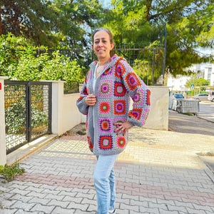 May include: A gray crochet cardigan with a colorful granny square pattern. The cardigan has a hood and a zipper closure. The model is wearing blue jeans and white sneakers.