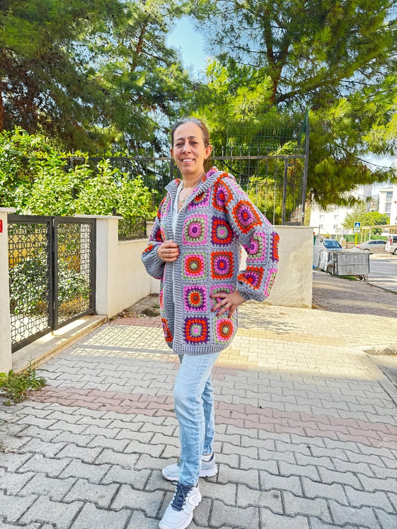 May include: A gray crochet cardigan with a colorful granny square pattern. The cardigan has a hood and a zipper closure. The model is wearing blue jeans and white sneakers.