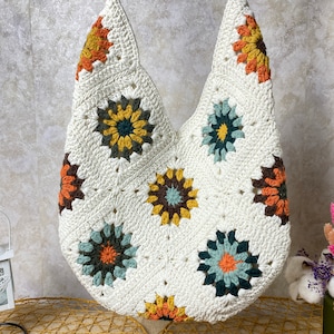 Op de afbeelding: Witte gehaakte tote bag met kleurrijke bloemenmotieven. De tas heeft een brede, open bovenkant en een enkele band.