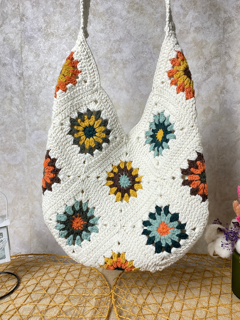 Op de afbeelding: Witte gehaakte tote bag met kleurrijke bloemenmotieven. De tas heeft een brede, open bovenkant en een enkele band.
