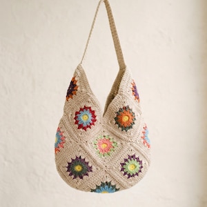May include: A beige crochet tote bag with a long strap. The bag features colourful floral designs in red, blue, orange, green, and purple. The bag is made of a soft, textured yarn.