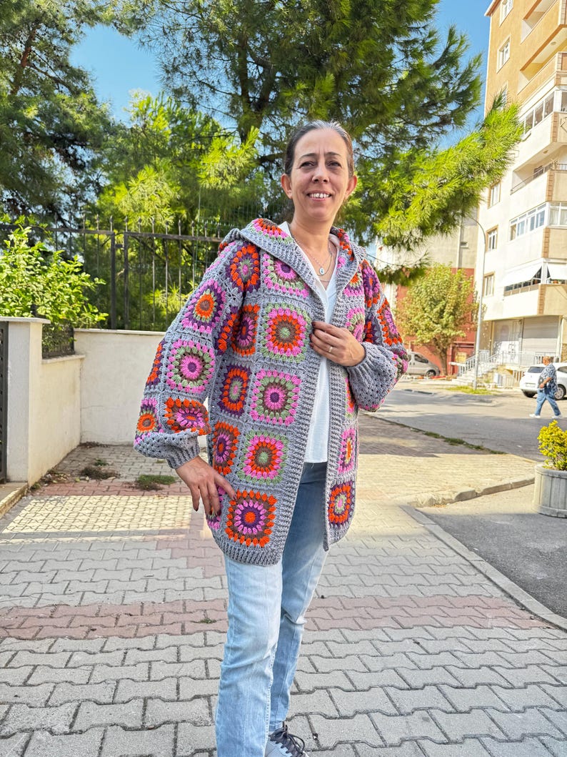 May include: A gray crochet cardigan with a hood. The cardigan has a colorful geometric pattern with pink, orange, green, and purple accents. The cardigan is worn over a white shirt and blue jeans.