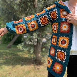 K&ouml;nnte beinhalten: Eine tealblaue geh&auml;kelte Strickjacke mit Kapuze, die ein farbenfrohes geometrisches Muster in Orange, Gelb und Wei&szlig; aufweist. Die Strickjacke ist aus einem weichen, strukturierten Garn gefertigt.