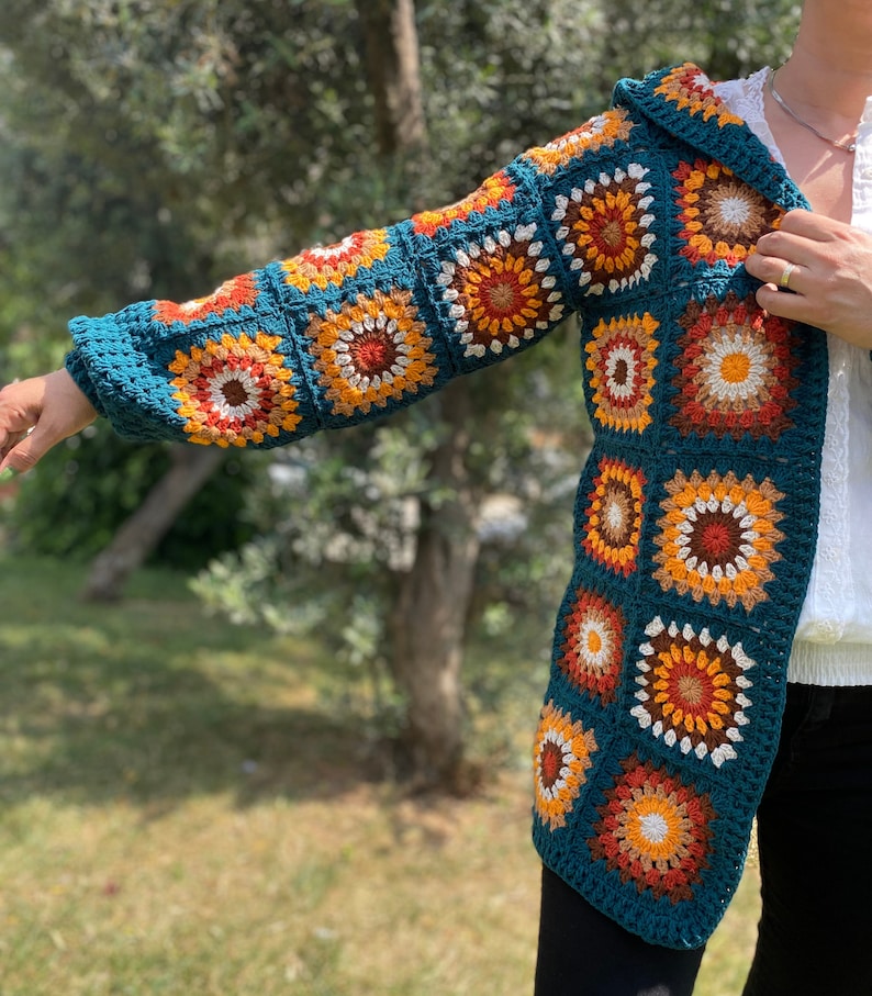 K&ouml;nnte beinhalten: Eine tealblaue geh&auml;kelte Strickjacke mit Kapuze, die ein farbenfrohes geometrisches Muster in Orange, Gelb und Wei&szlig; aufweist. Die Strickjacke ist aus einem weichen, strukturierten Garn gefertigt.
