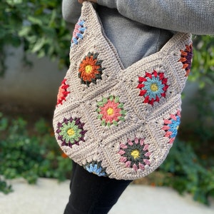May include: A beige crochet shoulder bag with colourful floral squares. The bag has a large, open design with a strap for carrying over the shoulder. The floral squares are in shades of orange, pink, blue, and green.