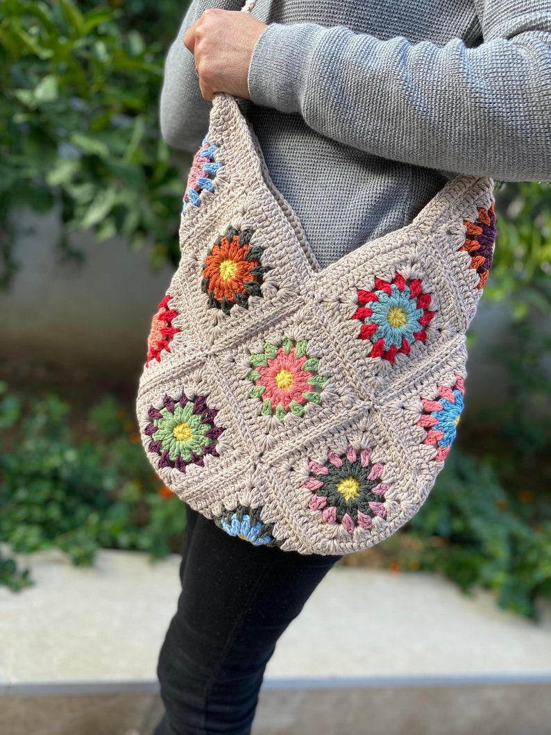May include: A beige crochet shoulder bag with colourful floral squares. The bag has a large, open design with a strap for carrying over the shoulder. The floral squares are in shades of orange, pink, blue, and green.