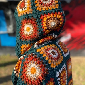 May include: A green crocheted hooded cardigan with a colorful geometric pattern. The cardigan is made with a variety of shades of brown, orange, yellow, and white.