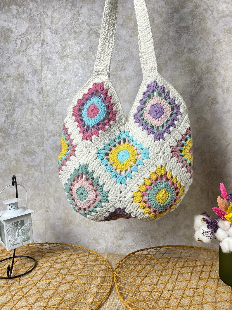 May include: A white crocheted tote bag with colorful floral designs. The bag has a wide, open top and two long straps.