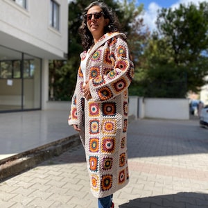 Puede incluir: Abrigo largo de ganchillo con capucha y dise&ntilde;o patchwork. El abrigo presenta cuadrados en tonos beige, naranja, amarillo, morado y verde. Se lleva sobre una camiseta blanca y vaqueros azules. El abrigo tiene mangas largas y un corte holgado.