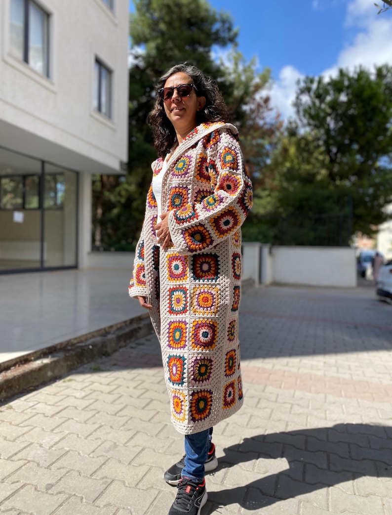 Puede incluir: Abrigo largo de ganchillo con capucha y dise&ntilde;o patchwork. El abrigo presenta cuadrados en tonos beige, naranja, amarillo, morado y verde. Se lleva sobre una camiseta blanca y vaqueros azules. El abrigo tiene mangas largas y un corte holgado.