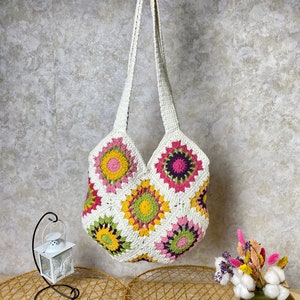 May include: White crochet tote bag with a colorful floral pattern. The bag has a long strap and is made of cotton yarn.
