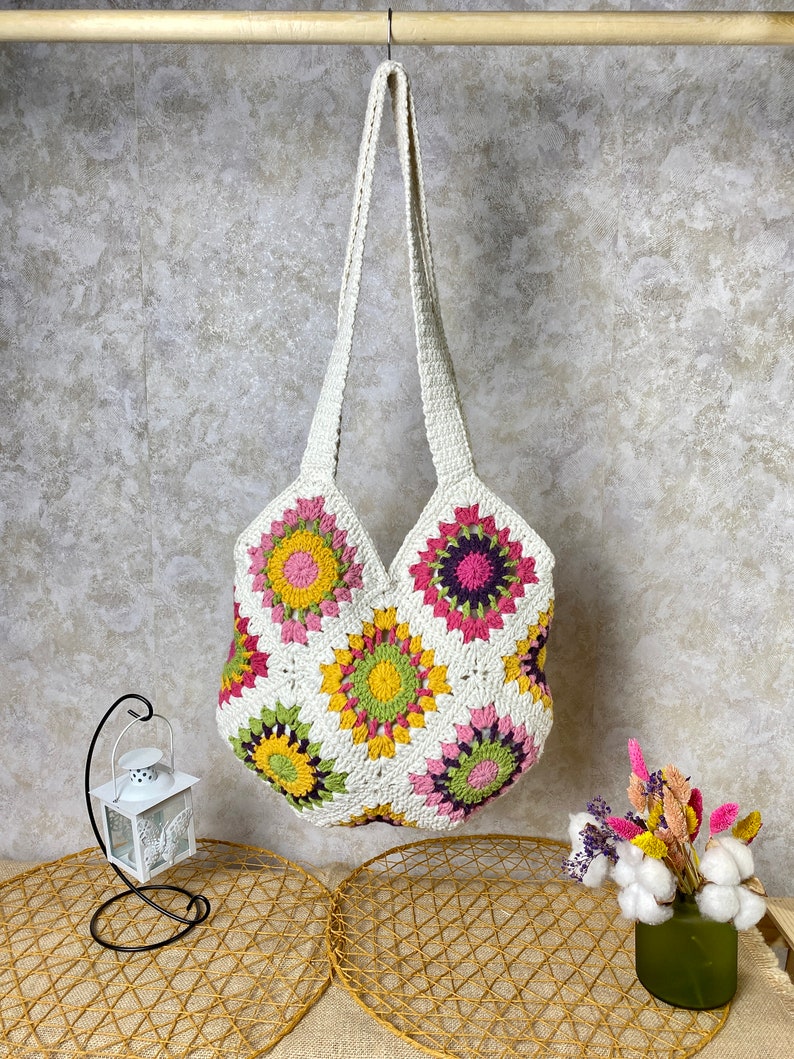 May include: White crochet tote bag with a colorful floral pattern. The bag has a long strap and is made of cotton yarn.