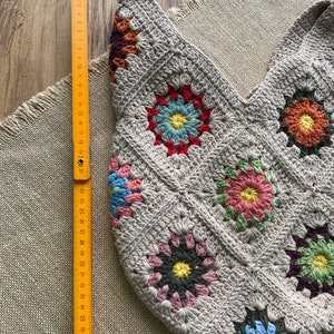 May include: A beige crocheted tote bag with a floral pattern. The bag is made of squares with colorful flower designs. A wooden ruler shows the bag is approximately 40 cm wide.