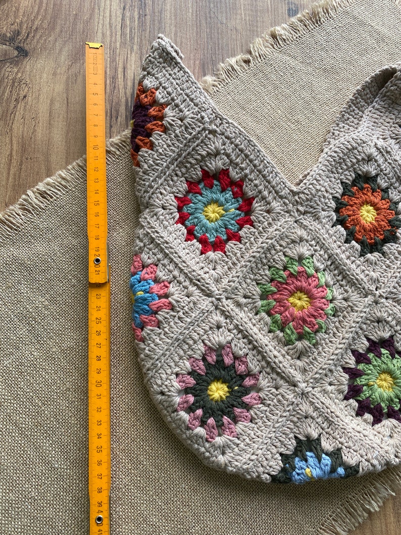 May include: A beige crocheted tote bag with a floral pattern. The bag is made of squares with colorful flower designs. A wooden ruler shows the bag is approximately 40 cm wide.