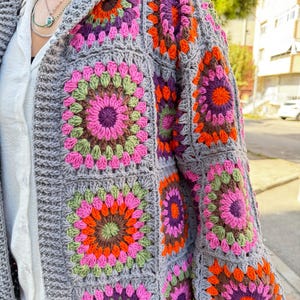 May include: A gray crochet cardigan with a hood, featuring colorful granny square patterns in shades of pink, orange, green, and purple.